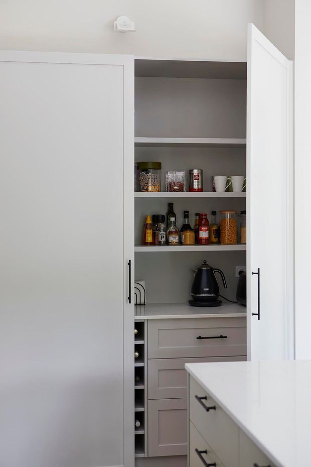A compact built-in kitchen pantry with white shaker-style doors, open grey shelving, white benchtop, drawers, black hardware, and neatly arranged food containers, bottles, cups, and a black electric kettle. Part of the kitchen island is visible in the foreground.