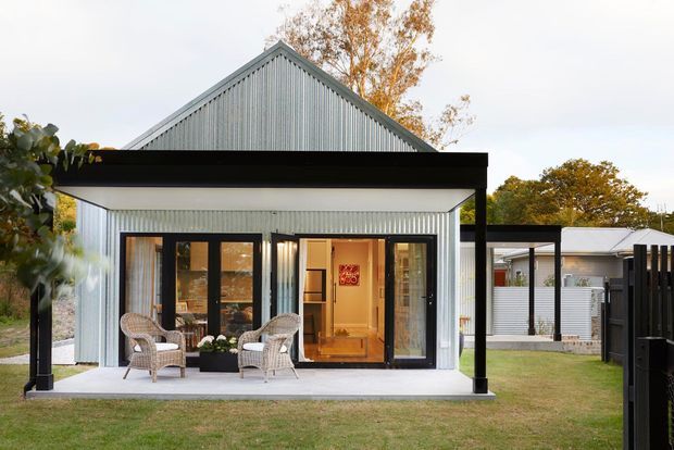 Exterior view of a compact single-storey gable-roofed dwelling with vertically corrugated light metal cladding, a deep black-framed covered porch, and black-framed glazed entry doors. Wicker chairs and small planters sit on the concrete porch, with a lawn and neighbouring outbuilding visible around the property.