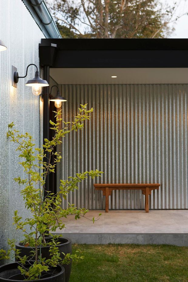 A covered outdoor patio or entry courtyard with corrugated metal walls, a black steel-framed canopy, concrete paving, wall-mounted gooseneck lights, potted plants, and a simple timber console table. The space has an industrial yet warm and inviting character, opening onto a small grassed garden area.