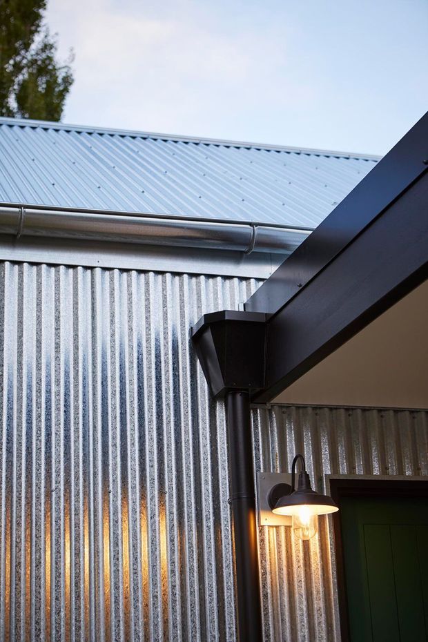 Close-up architectural detail of a corrugated metal-clad exterior wall beneath a pale corrugated metal roof, with visible guttering, dark structural eaves, downpipe, and a warm wall-mounted exterior lantern beside a dark green door. The image focuses on the building envelope and covered entry detail.