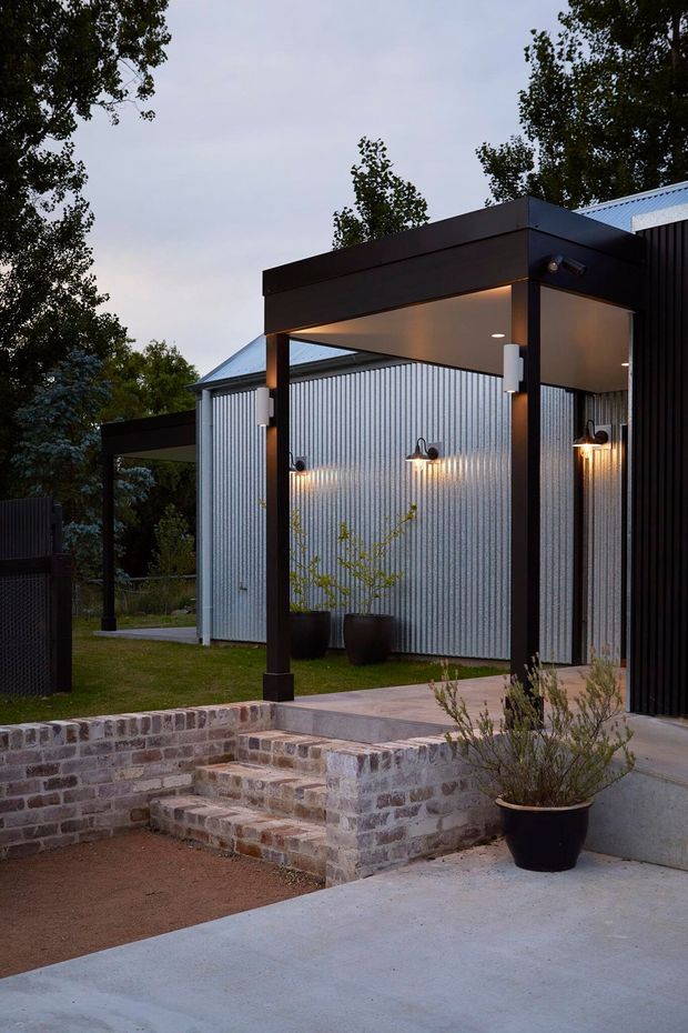 Twilight exterior view of a contemporary building with a covered entry canopy, black structural framing, corrugated metal wall cladding, and dark vertical cladding. Brick retaining walls and steps lead up from a concrete path, with integrated wall lighting, potted plants, and landscaped surroundings.