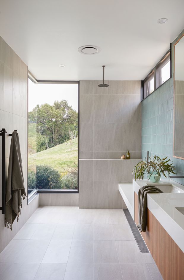 A bright contemporary bathroom with a walk-in shower, large-format pale grey wall and floor tiles, a freestanding or integrated white bathtub, and a timber-veneered vanity. Full-height glazing opens to a leafy sloping landscape, while pale blue-green vertical tiles form a feature wall. Minimal black-framed detailing, a rainfall shower, and soft natural light create a calm, refined atmosphere.