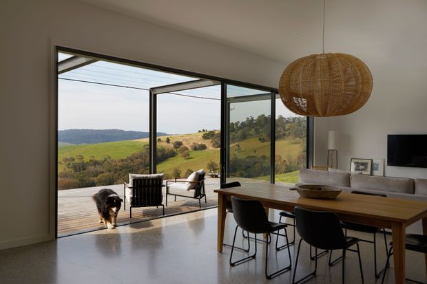 A contemporary open-plan dining and living area with a large timber dining table, dark dining chairs, a neutral sofa, and a woven pendant light. Full-height black-framed glazing and open glass doors connect the room to a terrace, overlooking a rolling rural landscape. The interior is bright and minimally furnished, with polished light flooring and warm natural accents.