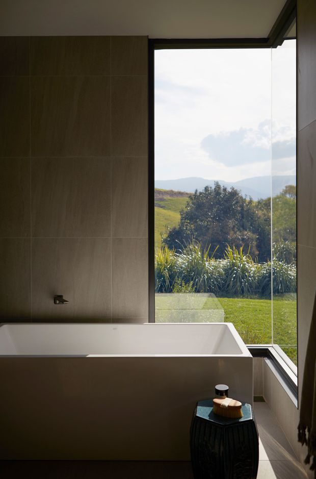 A refined contemporary bathroom with a freestanding white bathtub positioned beside full-height glazing overlooking a landscaped lawn and rolling hills. Large taupe-brown wall tiles create a calm, earthy backdrop, while dark window frames, a small black side table, and a folded textile add contrast. The composition is warm, minimalist, and strongly connected to the surrounding landscape.