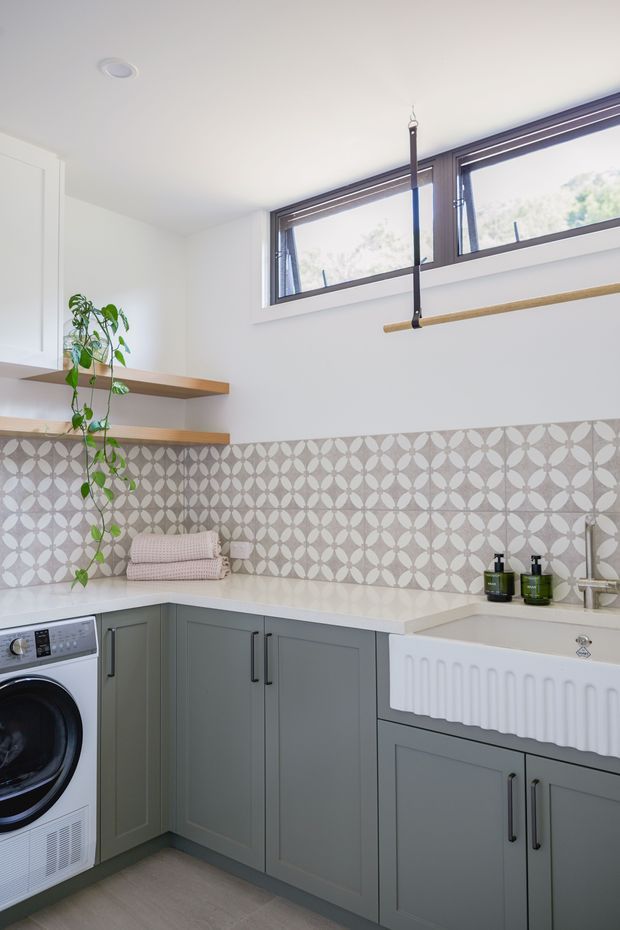 Bright, compact laundry room with grey-green shaker-style cabinetry, white benchtops, a large white farmhouse utility sink, and a patterned grey and white tiled splashback. Timber open shelving, a hanging rail, front-loading washing machine, trailing plant, and high horizontal windows complete the functional interior.