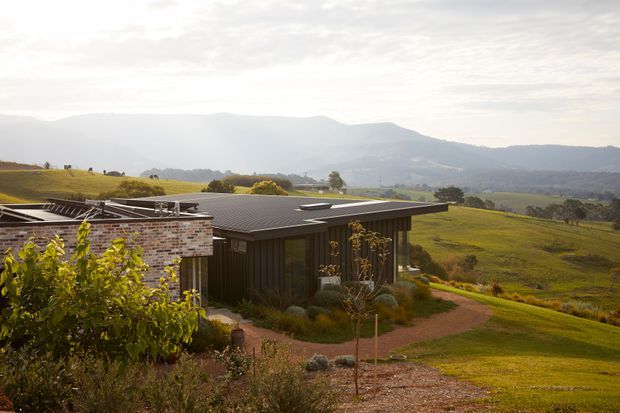 A low-profile contemporary rural house set on a gently sloping landscaped site, with dark vertical cladding, a broad shallow roof, and a brick-clad wing. The home overlooks expansive green pasture and a mountainous landscape, with planted garden beds and a curving path in the foreground. Warm, soft daylight creates a calm pastoral atmosphere.
