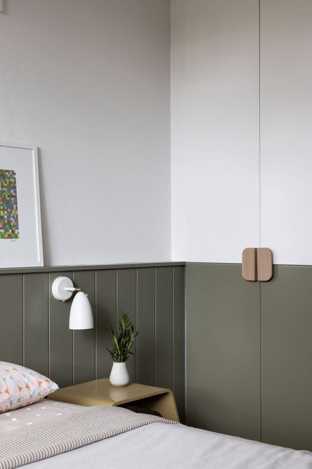 A calm, contemporary bedroom corner with a neatly made bed, dark olive-green vertical wall panelling below white painted walls, a slim integrated bedside table, white wall-mounted lamp, small potted plant, framed artwork, and a minimalist wooden wall hook or handle detail. The palette is muted and softly lit, with understated Scandinavian-inspired styling.