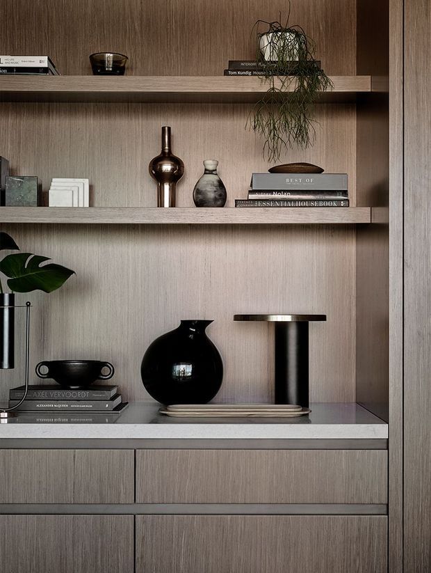 Close-up interior detail of a refined built-in timber-veneer shelving and cabinetry unit. Open shelves display books, ceramics, sculptural vases, a table lamp, and a trailing plant, with lower drawers providing concealed storage. The composition is warm, muted, and carefully styled.