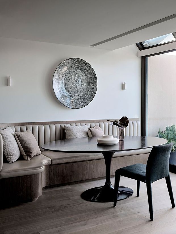 A refined dining nook with a curved upholstered banquette, round pedestal dining table, and upholstered dining chair. The space features pale neutral walls, dark timber flooring, sculptural wall art, integrated linear ceiling detailing, wall sconces, and abundant natural light from a large window and skylight.