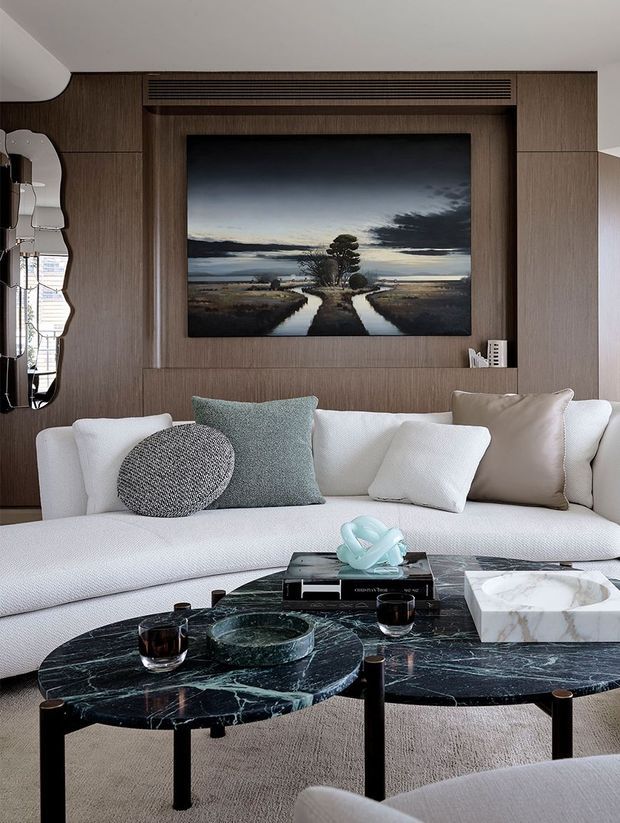 A refined contemporary living room centered on a large white sectional sofa, layered cushions, and low round coffee tables with dark veined stone tops and black legs. The room features a warm dark timber wall with integrated slatted ventilation above a wall-mounted artwork or screen, a sculptural irregular mirror, soft carpeting, and restrained decorative objects. The palette is neutral and sophisticated with white upholstery, dark wood, black stone, and muted blue-green accents.