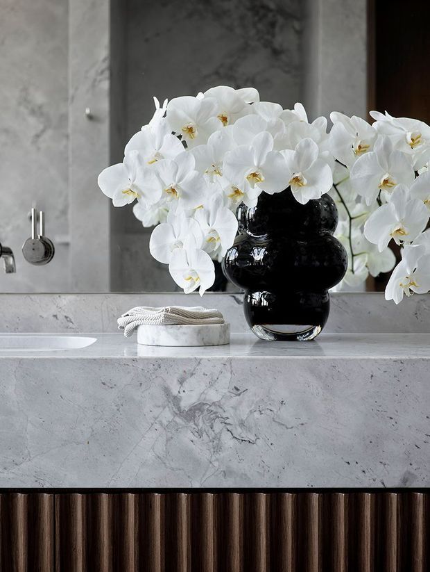 Close-up view of a refined bathroom vanity featuring a pale grey marble or stone benchtop and splashback, integrated white basin, dark fluted timber cabinetry, chrome fixtures, and a glossy black sculptural vase filled with white orchids. The styling is luxurious and monochromatic with strong veining and reflective surfaces.