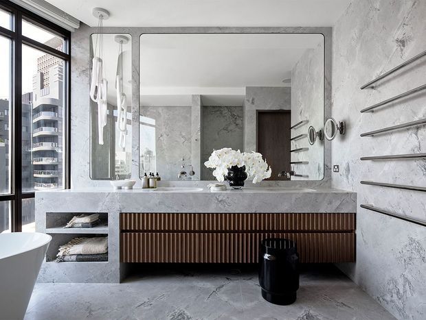 A luxurious contemporary bathroom with a double vanity beneath a large rounded-rectangle mirror. The room features grey veined stone or concrete-look wall and floor finishes, a pale stone benchtop, dark vertically fluted timber vanity cabinetry, exposed metal towel rails, and a freestanding white bathtub at the edge of the frame. Large black-framed windows provide an urban view, while sculptural pendant lights and monochrome accessories add refinement.