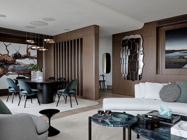 A refined open-plan living and dining interior with warm timber wall panelling, a sculptural black dining table and upholstered dining chairs, a large white sectional sofa, and layered contemporary lighting. Decorative artwork, a statement mirrored wall piece, patterned marble coffee tables, and neutral furnishings create an elegant, richly textured space.