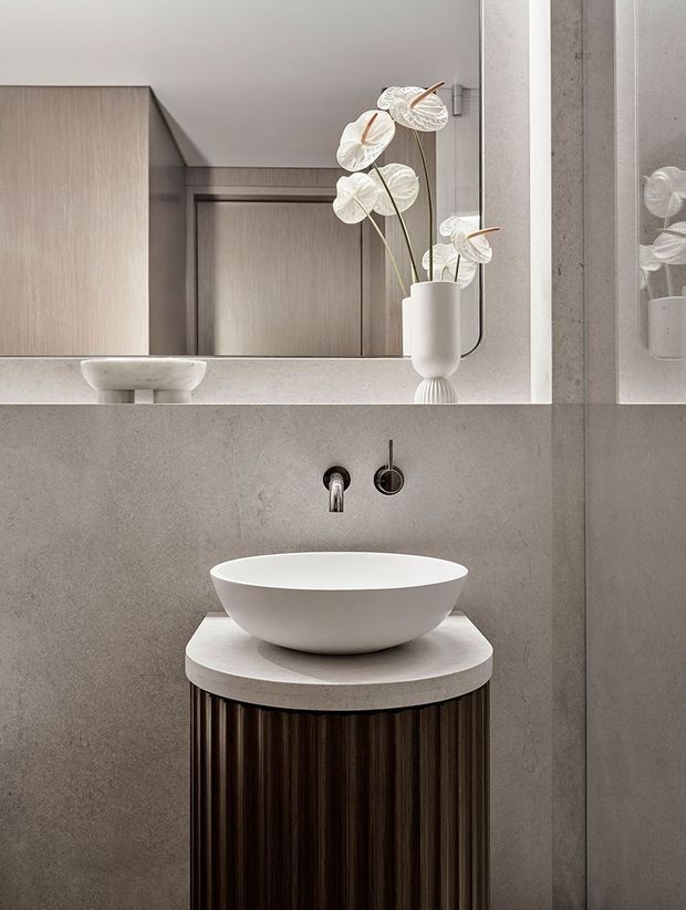 A refined contemporary bathroom vanity in a compact space. A white round vessel basin sits on a cylindrical dark fluted timber vanity with a pale stone benchtop. The walls are finished in warm grey textured material, with minimalist wall-mounted plumbing and a large mirror reflecting cabinetry and decorative white flowers. The palette is neutral, warm, and understated.
