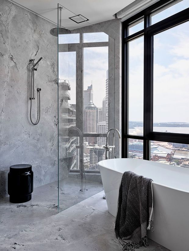 A high-rise bathroom with a frameless glass walk-in shower, freestanding white bathtub, wall-mounted shower fixtures, and a black freestanding stool or side table. Grey textured concrete-look wall and floor finishes contrast with black window frames and expansive city and waterfront views.