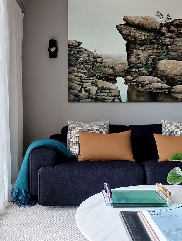 A styled contemporary living room vignette featuring a deep navy sofa with camel and patterned cushions, a teal throw, pale grey walls, sheer curtains, and a large landscape artwork depicting rocky formations. A round white marble coffee table with books and magazines occupies the foreground, while a black geometric wall sconce provides a small architectural accent.