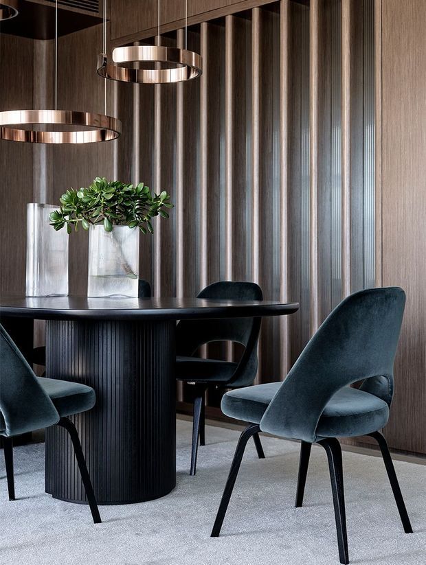 Sophisticated dining area with a round black dining table, sculptural upholstered charcoal dining chairs, layered circular pendant lights, and a tall glass planter. The room features dark timber wall panelling with vertical fluted and metallic detailing, creating a refined, dramatic contemporary interior.