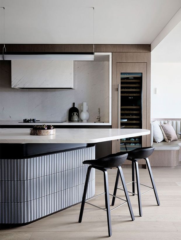 A refined contemporary kitchen with a curved island and integrated seating. The island features a pale stone benchtop, dark vertically fluted cabinetry, and black bar stools. Behind it is a full-height marble-look splashback, dark timber cabinetry, a concealed rangehood, cooktop, sink, and built-in wine fridge. A banquette seating area is visible to the right, with light timber flooring and a restrained neutral palette.