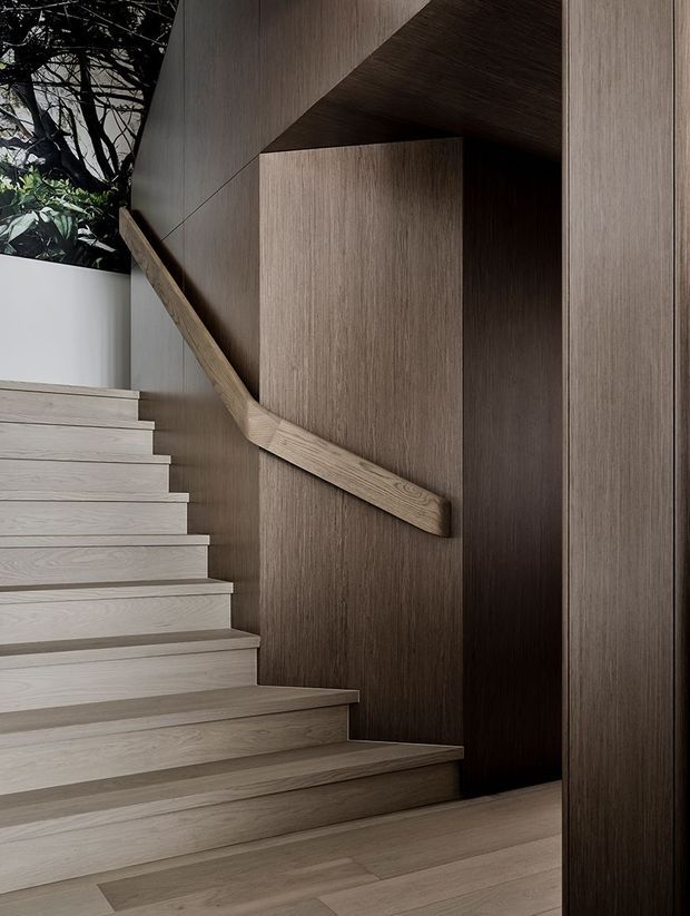 A refined contemporary stairway clad in pale and medium-toned timber, with broad timber treads, a sculptural angled timber handrail, and dark vertical wood-panelled walls. The stair turns through a carefully detailed, minimalist enclosure with a small glimpse of planting visible beyond.