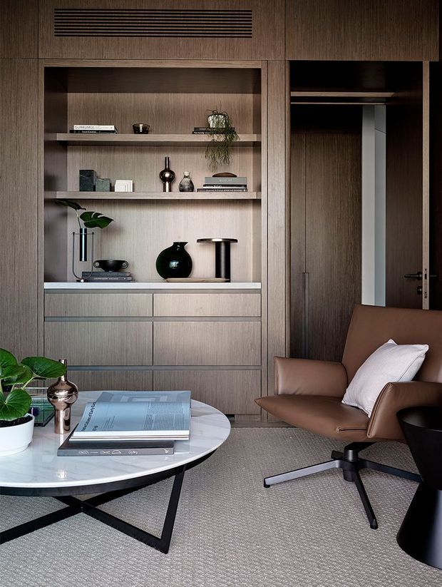 A refined bedroom interior featuring full-height dark timber built-in cabinetry with open display shelving, drawers, and a concealed wardrobe. A round marble coffee table, upholstered swivel lounge chair, black sculptural side table, indoor plants, books, and decorative objects create a warm, sophisticated reading or sitting area. The palette is dominated by dark wood, taupe upholstery, white marble, and soft grey carpeting.
