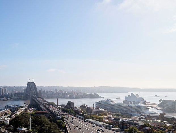 Wide elevated cityscape view over Sydney Harbour, showing the Harbour Bridge, Opera House, cruise ships, dense urban buildings, and multilane road infrastructure under a bright blue sky.