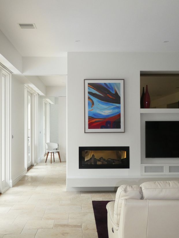 Bright contemporary living room with a built-in linear fireplace and wall-mounted television set within white cabinetry. A colourful abstract artwork hangs above the fireplace, while a white leather sofa, pale stone-look flooring, and a glazed corridor with a small chair are visible. The room has clean white walls, recessed lighting, and abundant natural light.