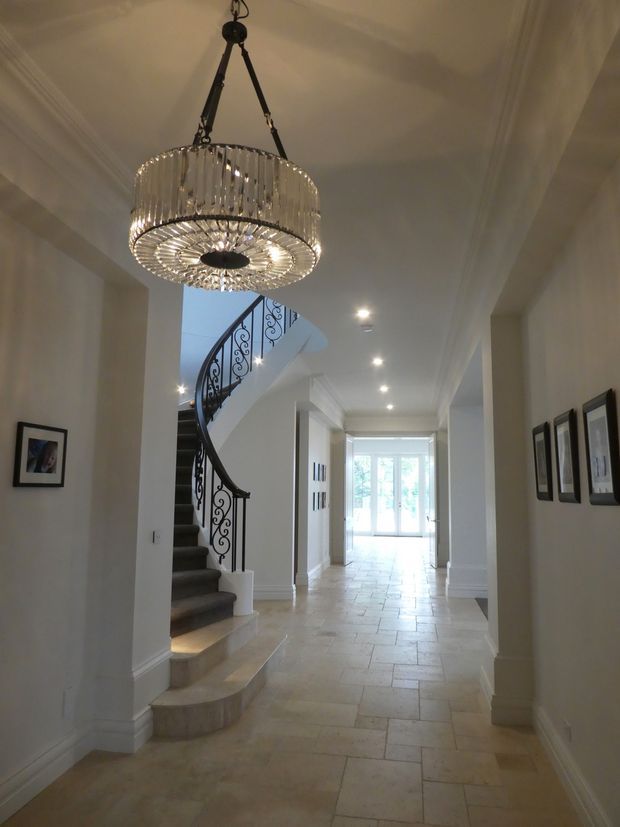 Elegant residential entrance hallway with pale stone tile flooring, white walls and detailed cornices, a curved staircase with ornate black wrought-iron balustrade, framed artwork, recessed lighting, and a large crystal chandelier. Bright glazing at the far end creates an airy, refined atmosphere.