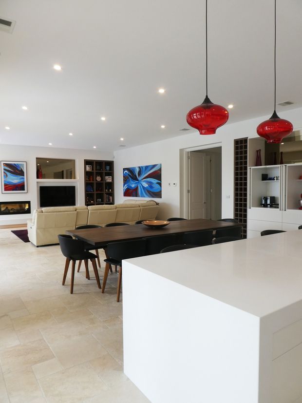 Open-plan contemporary interior viewed from the kitchen, with a large white island in the foreground, a dark timber dining table with black chairs, and a living area beyond. The space features pale tiled flooring, white walls and cabinetry, recessed ceiling lights, red glass pendant lights, built-in shelving, a television, and colourful abstract artwork.