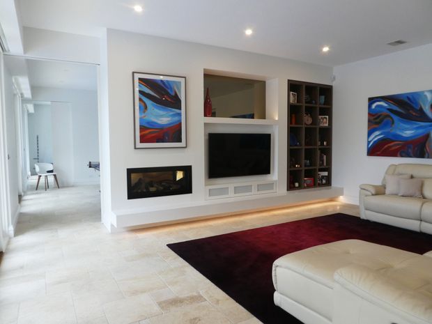 Bright contemporary living room with a built-in media wall, recessed fireplace, open display shelving, and integrated LED lighting. Pale tiled flooring and white walls are paired with cream leather seating, a dark burgundy rug, and colourful abstract artwork. A hallway and small workspace are visible to the left.