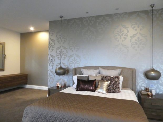 A refined bedroom with a large upholstered bed, layered neutral bedding and decorative cushions, set against a silver-grey damask feature wall. Two sculptural pendant lights hang symmetrically over dark bedside tables, with a low timber console along the side wall. The room has warm, muted finishes and polished contemporary detailing.