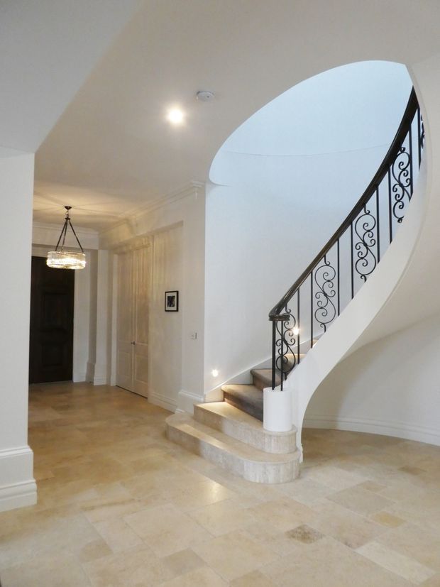 Elegant residential entry hall with a sweeping curved staircase, ornate black wrought-iron balustrade, pale stone flooring and stair treads, white plaster walls with classical mouldings, paneled doors, and a decorative pendant light.