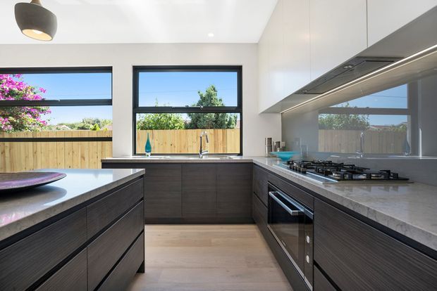 Contemporary galley-style kitchen with dark wood-grain cabinetry, pale engineered-stone benchtops, integrated stainless-steel appliances, a grey splashback, and light timber flooring. Large black-framed windows overlook a fenced garden, while under-cabinet lighting and a sculptural pendant provide illumination.