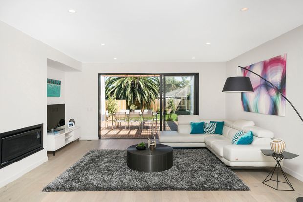 Bright contemporary living room with a white sectional sofa, black round coffee table, grey shag rug, wall-mounted fireplace, abstract colourful artwork, and large sliding glass doors opening to a furnished outdoor terrace and garden. Recessed ceiling lighting and minimalist built-in media cabinetry complete the space.