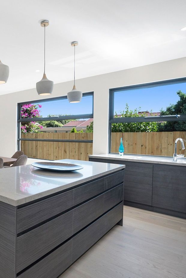 Bright contemporary kitchen featuring a large central island with a pale stone benchtop and dark horizontal-grain built-in cabinetry. Matching cabinetry runs beneath expansive dark-framed windows, with pendant lights above the island, a stainless-steel sink and tap, and light timber-look flooring. The windows overlook a fenced suburban garden.