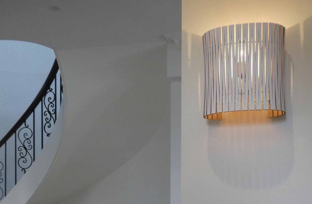 Interior architectural detail showing a curved stairwell with ornate black metal balustrade on the left and a sculptural wall-mounted light fixture made of vertical slats on the right. The space has white painted walls and a clean, refined appearance.