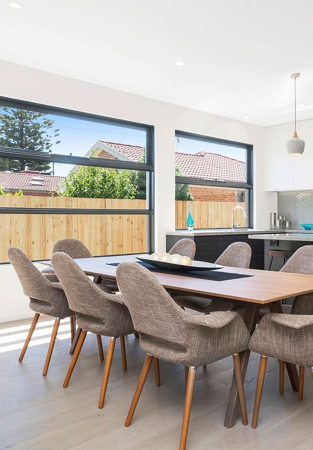 Bright contemporary dining area with a large timber dining table surrounded by upholstered dining chairs. The room opens visually to a compact kitchen with dark cabinetry, a light benchtop, stainless-steel sink and tapware, and a pendant light. Large black-framed windows provide abundant daylight and overlook a timber fence and neighbouring tiled roofs.