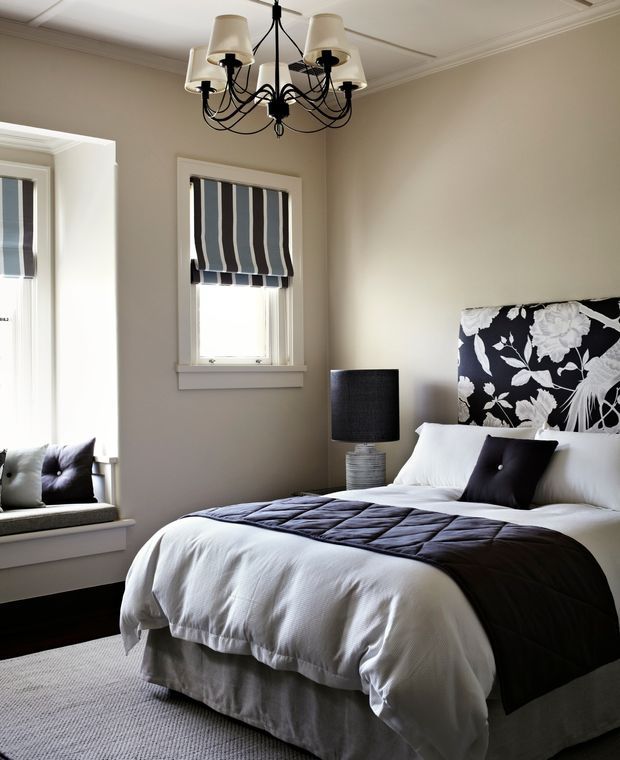 A refined bedroom with a white upholstered bed dressed in white bedding and a dark charcoal quilted runner. A bold black-and-white floral upholstered headboard forms the main feature, complemented by dark accent cushions and a black bedside lamp. The room has warm neutral walls, white architectural trim and ceiling moulding, striped Roman blinds, a window seat with cushions, dark timber flooring, and a pale textured rug. A traditional multi-arm chandelier provides decorative lighting.