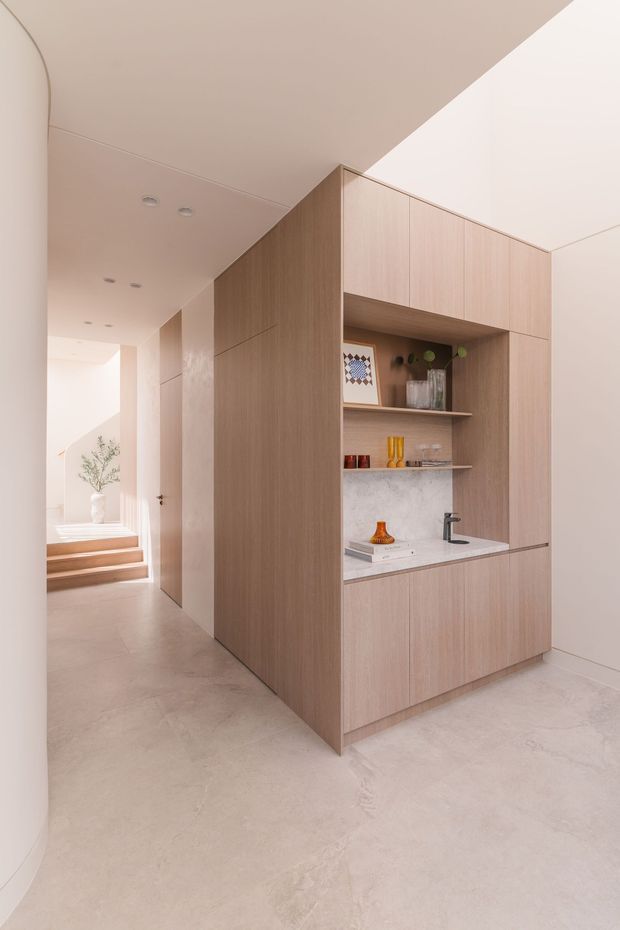 A minimalist open-plan interior hallway featuring a full-height light timber joinery wall with integrated cabinetry, open display shelves, and a compact stone-topped wet bar or kitchenette with a black tap. Pale polished flooring, warm off-white walls, recessed ceiling lights, and a softly lit stairway create a refined, neutral atmosphere.
