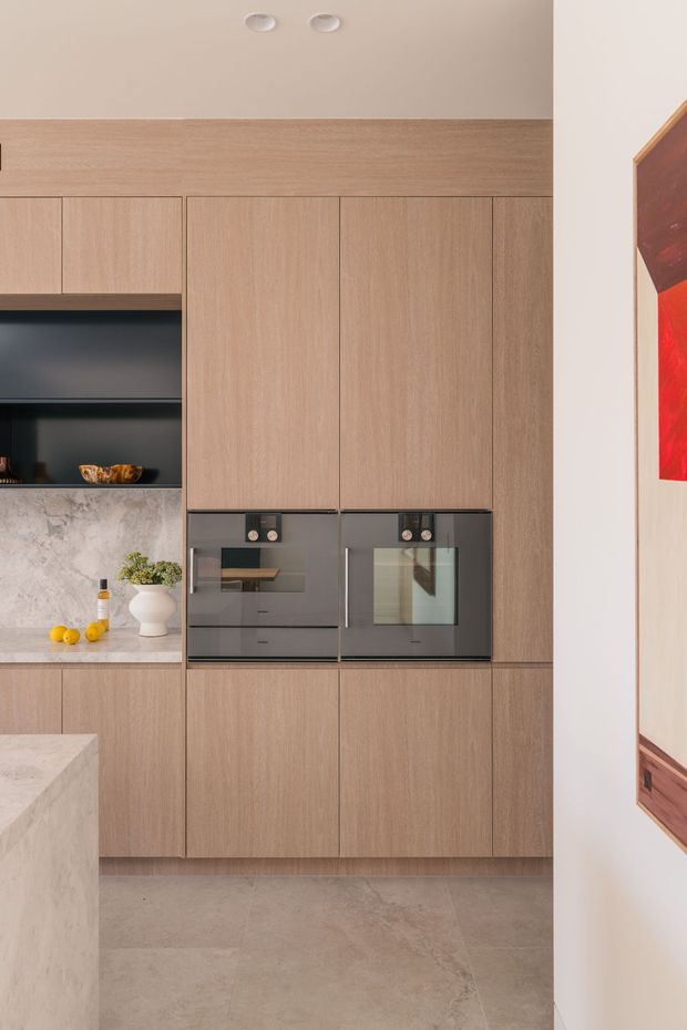 A refined contemporary kitchen featuring a full wall of light timber cabinetry, integrated dark glass ovens, a dark open display niche, and a pale veined stone benchtop and splashback. Large-format neutral stone flooring and recessed ceiling lights create a clean, warm, minimalist look. A partial island and framed artwork are visible at the edges.