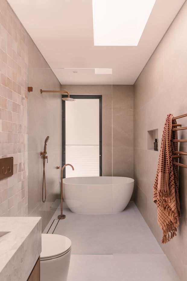 A narrow, refined bathroom featuring a freestanding white bathtub, wall-mounted shower, frameless glass shower partition, wall-hung toilet, large skylight, pale stone-look wall and floor finishes, and warm bronze plumbing and heated towel rail. Soft blush-beige tones create a calm, spa-like atmosphere.