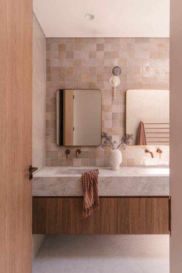A refined contemporary bathroom viewed through an open timber door. The room features a floating dark timber vanity with a pale stone benchtop, warm pink-beige square tiled walls, two rounded-rectangle mirrors, wall-mounted copper-toned tapware, and soft sculptural lighting. A neutral woven towel and ceramic vase add warmth and texture.
