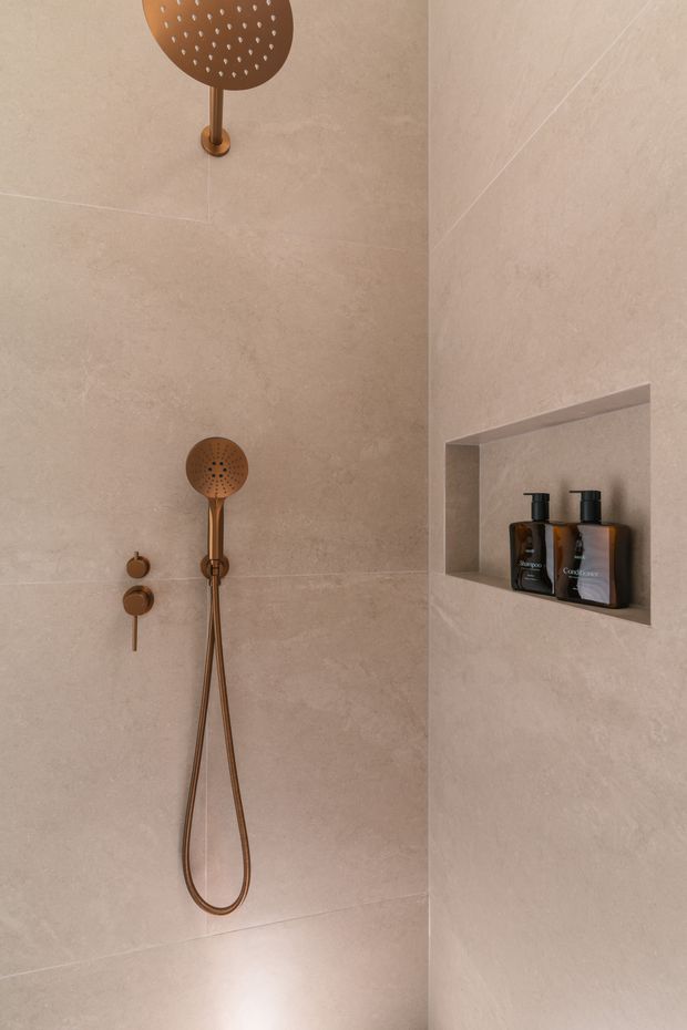 Close-up view of a refined walk-in shower clad in large warm beige stone-look tiles. The shower features brushed bronze or copper-toned fixtures, including a round overhead rain shower, handheld shower, controls, and hose. A recessed wall niche holds dark soap and shampoo dispensers, creating a clean, minimalist and warm bathroom interior.
