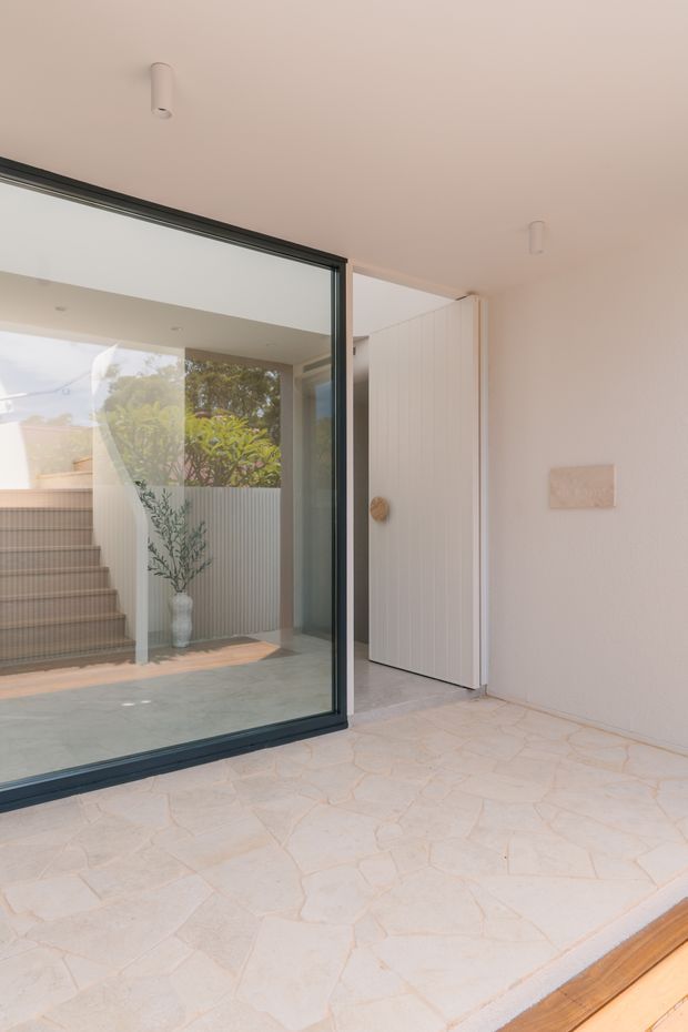 A bright, minimalist residential entryway or transitional foyer with pale irregular stone or tile flooring, white textured walls and ceiling, a full-height black-framed glass partition, and a white vertical-panel door. Through the glazing, a small internal courtyard or stair landing is visible with tiled stairs, ribbed wall cladding, and a potted plant. Warm timber edging appears at the foreground threshold.
