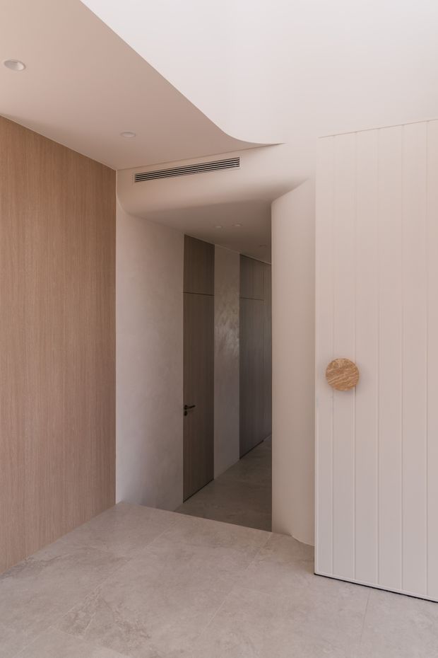 A minimalist residential hallway or entry area with warm neutral finishes. Large vertical timber wall panelling lines the left side, while pale textured wall panelling and a tall white built-in cabinet occupy the right. A narrow corridor with flush timber doors extends into the background. The floor is finished in large pale stone-look tiles, and the sculptural curved ceiling incorporates recessed downlights and a linear air-conditioning vent.