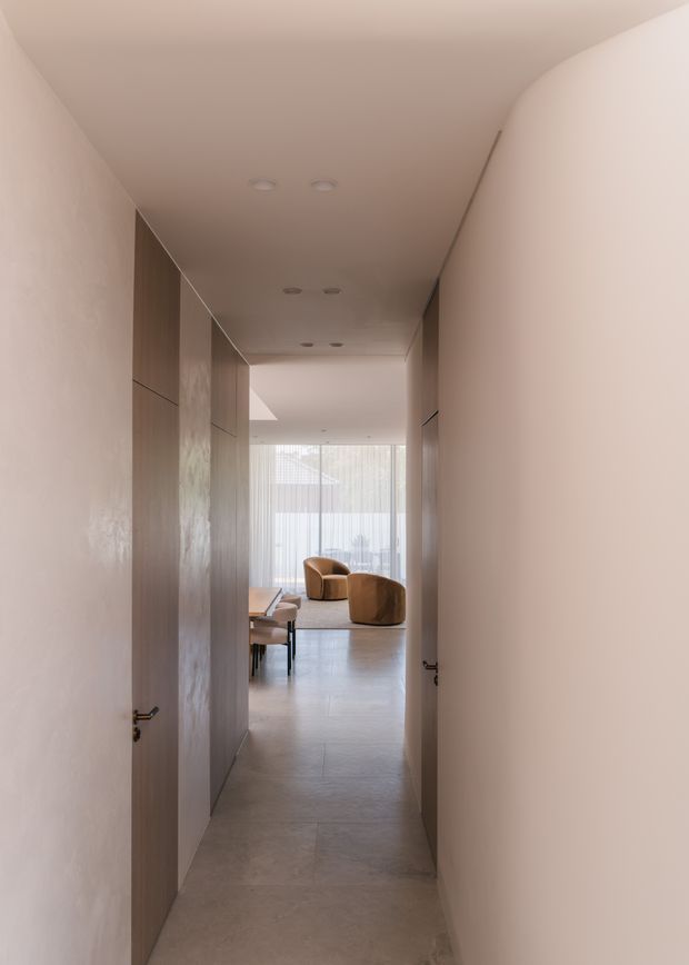 A minimalist interior hallway viewed through partially open doors, leading toward a bright living area with floor-to-ceiling glazing and two rounded brown lounge chairs. The corridor has smooth warm-white walls, flush timber-toned built-in doors or cabinetry, a pale stone or concrete-look floor, and recessed ceiling lighting.