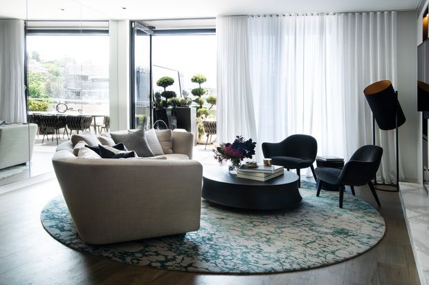 A bright, refined contemporary living room with a large curved sectional sofa, two dark lounge chairs, a low round coffee table, and a patterned area rug. Full-height sheer curtains and floor-to-ceiling glazing open to an outdoor terrace, while a dining area is visible beyond. The space has light timber flooring, pale walls, sculptural furnishings, and abundant natural light.