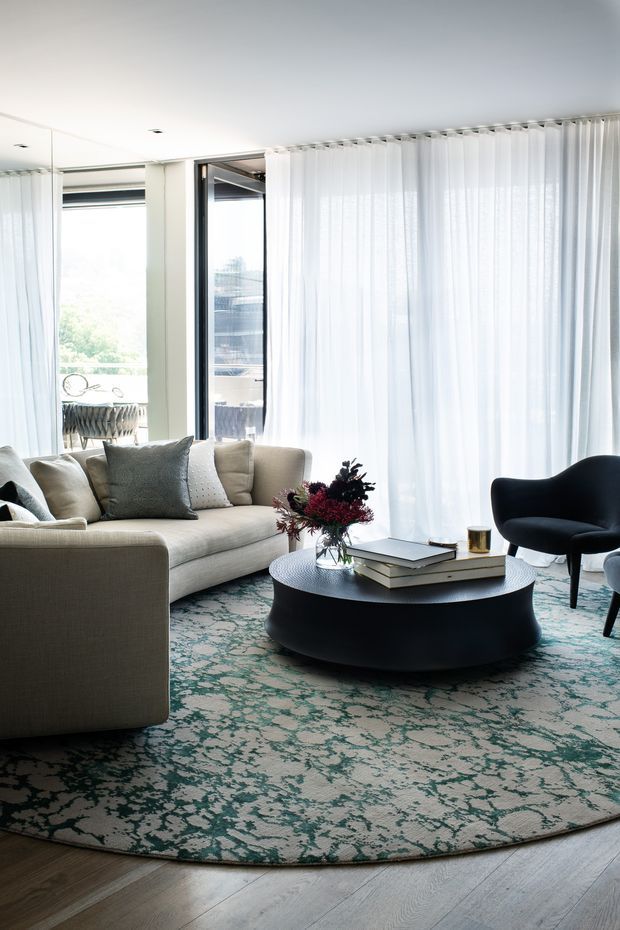 Bright contemporary living room with floor-to-ceiling sheer curtains and glazing, a large light upholstered sectional sofa, sculptural black lounge chair, round black coffee table, patterned teal and cream area rug, and floral arrangement. The room has a refined, softly layered neutral palette with strong natural light and views through the windows.