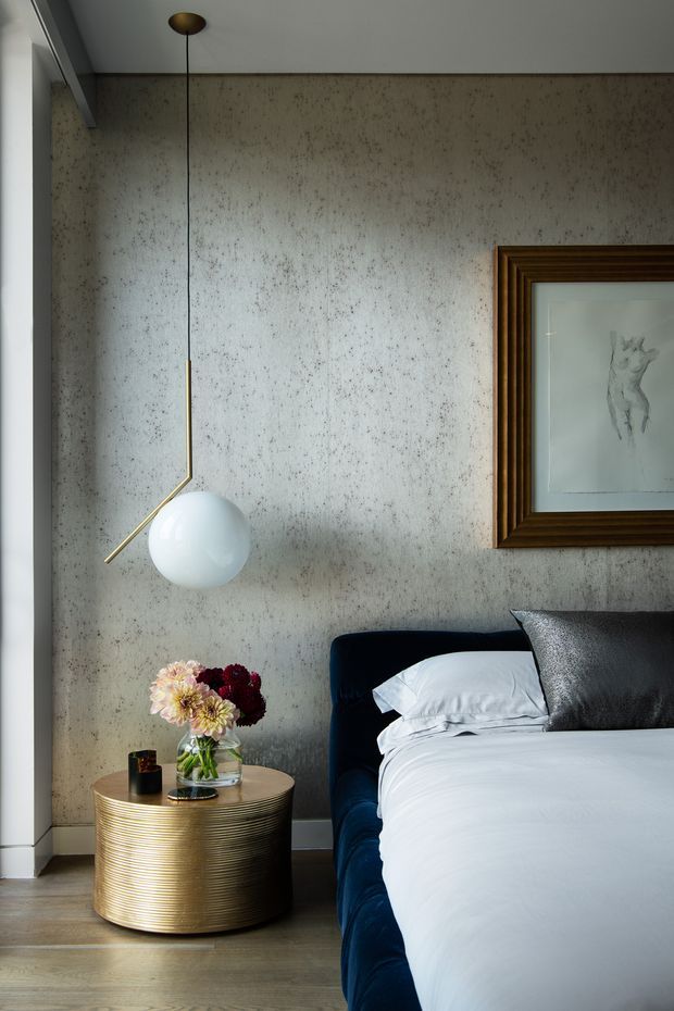 A refined bedroom corner featuring a deep navy upholstered bed, crisp white bedding, a cylindrical ribbed metallic-gold bedside table, and a sculptural globe pendant light. The room has a mottled textured grey wall, pale timber flooring, and a large dark timber-framed artwork, creating a moody yet luxurious contemporary setting.