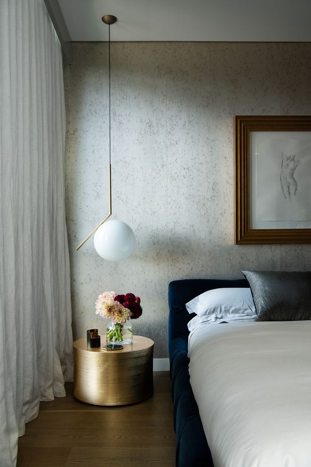A refined bedroom corner featuring a deep navy upholstered bed, crisp white bedding, dark flooring, a ribbed metallic-gold cylindrical bedside table, and a sculptural spherical pendant light with brass detailing. Full-height sheer curtains soften the window wall, while a textured grey plaster or concrete-look feature wall and framed artwork add visual depth.