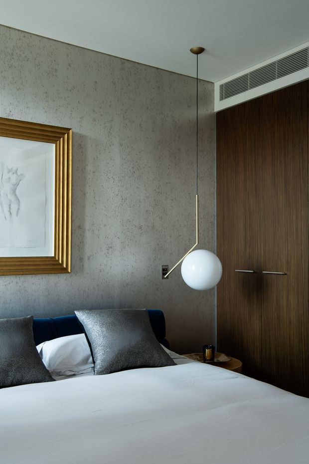 A sophisticated bedroom interior featuring a neatly made bed with white bedding, dark charcoal textured cushions, and a deep navy upholstered headboard. The room has a mottled grey textured wall finish, dark timber built-in wardrobe doors, a brass-framed artwork, and a sculptural spherical pendant light beside the bed. The overall composition is refined, moody, and contemporary with warm timber and metallic accents.